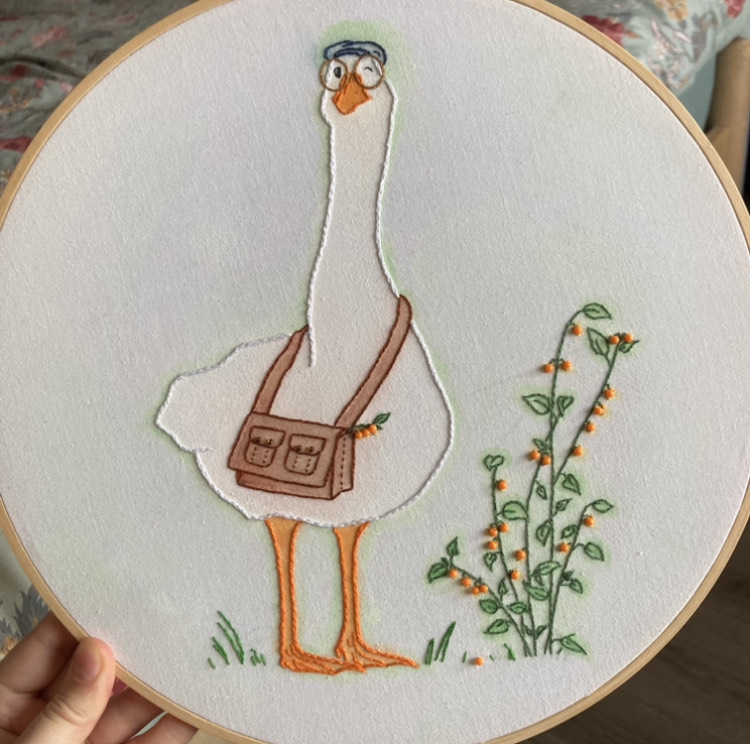 embroidered goose on light green watercolour background, linen on wooden hoop. the goose is wearing a messenger bag, round eye glasses, and a newsboy cap. he is standing next to a gooseberry bush.